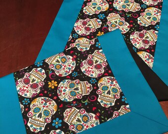 May include: Two decorative table runners. The center fabric features a repeating pattern of colorful sugar skulls on a black background. The edges are a solid turquoise blue. The runners are likely made of fabric.