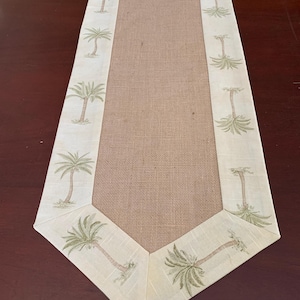Puede incluir: Un camino de mesa beige con un centro de arpillera y un borde de palmeras. El borde es de color beige claro con palmeras verdes.
