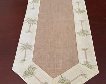 May include: A beige table runner with a burlap center and a border of palm trees. The border is a light beige color with green palm trees.
