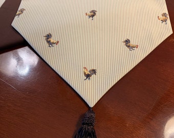 May include: A white and brown striped table runner with a black tassel. The runner features a repeating pattern of embroidered roosters.