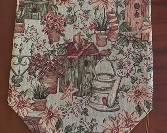 May include: A beige and brown table runner with a floral pattern featuring birdhouses, watering cans, and potted plants. The runner has a scalloped edge.