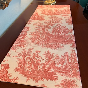 May include: A red and white toile table runner with a French country design featuring a pastoral scene with people and trees.