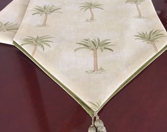 May include: A light green table runner with a repeating pattern of palm trees. The runner has a green border and is finished with two matching tassels. The fabric is a light beige color.