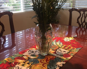 May include: A table runner with a skull and rose design. The runner is red, yellow, blue, and green. The text "Carpe Noctem" is printed on the runner.