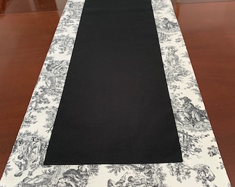 May include: A black and white table runner with a black center and a white border featuring a toile pattern.