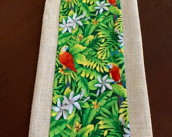 May include: A beige table runner with a green, yellow, and white tropical print featuring parrots and flowers. The runner has a burlap border.
