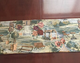 May include: A tapestry runner with a garden scene featuring a tennis court, a gazebo, a fountain, and flowers. The colors are green, brown, pink, and white.