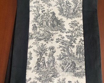 May include: A black and white toile table runner. The center features a detailed pastoral scene with figures, trees, and buildings. The edges are solid black, creating a framed effect. This runner is suitable for dining room decor.