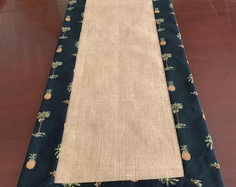 May include: A burlap table runner with a black fabric border featuring a pineapple pattern. The border is approximately 2 inches wide.