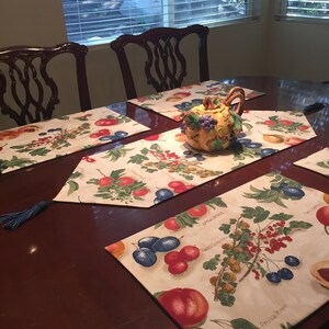 May include: Four placemats with a fruit and berry design, a teapot with a fruit and berry design, and a wooden table. The placemats are cream colored with a black border and feature a variety of fruits and berries, including plums, cherries, apples, and grapes. The teapot is yellow with a green handle and spout and is decorated with a variety of fruits and berries. The table is dark brown wood.