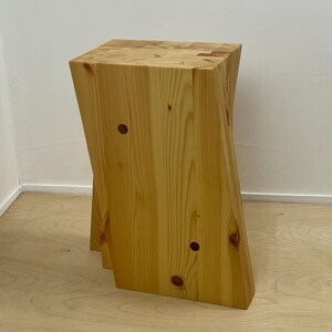 Op de afbeelding: Een lichtgekleurde houten kruk met een vierkante bovenkant en een licht schuin, asymmetrisch ontwerp. De kruk is gemaakt van massief hout, met zichtbare houtnerf en knopen. Het heeft een natuurlijke afwerking en een minimalistische esthetiek.