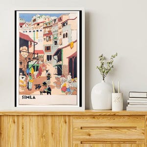 May include: A framed vintage travel poster for Simla, India, depicting a bustling street scene with colorful buildings and figures. The poster has a white border and is displayed on a wooden cabinet.