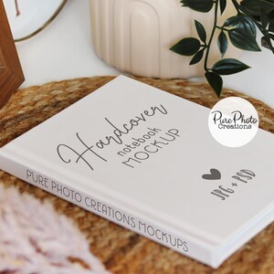Pode incluir: Maquete de caderno de capa dura branca com o texto "Hardcover notebook MOCKUP" e "PURE PHOTO CREATIONS MOCKUPS" na capa. O caderno est&aacute; sobre um tapete tecido marrom com uma pequena planta em vaso e um rel&oacute;gio branco no fundo.