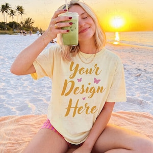 May include: A light yellow t-shirt with the text "Your Design Here" in gold script and pink butterflies. The person is holding a green iced drink with a straw. The background is a beach at sunset.