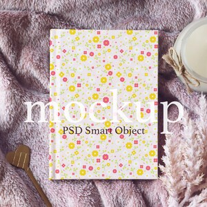 May include: A white notebook with a pink and yellow floral pattern. The notebook is lying on a pink fuzzy blanket with a candle and dried flowers.