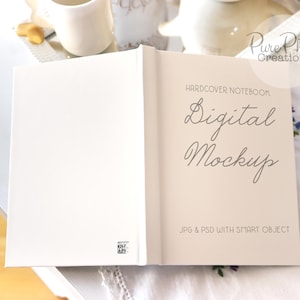 Puede incluir: Una maqueta de cuaderno de tapa dura blanca con el texto "HARDCOVER NOTEBOOK Digital Mockup JPG & PSD WITH SMART OBJECT" en la portada.