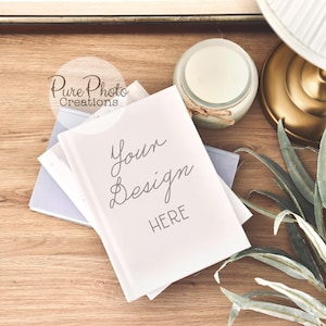 Puede incluir: Un libro blanco con el texto "Your Design Here" sobre una superficie de madera con una vela, una lámpara y una planta verde.