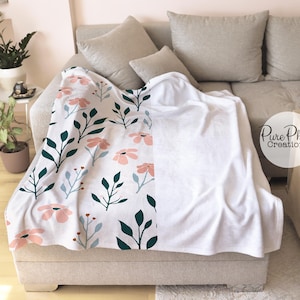 May include: A white fleece throw blanket with a floral pattern on one side. The pattern features pink flowers, green leaves, and white stems on a white background. The other side of the blanket is plain white.