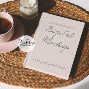 Puede incluir: Un cuaderno blanco de tapa dura con el texto "HARDCOVER NOTEBOOK Digital Mockup JPG & PSD WITH SMART OBJECT" en la portada. El cuaderno está sobre una estera de mimbre marrón.