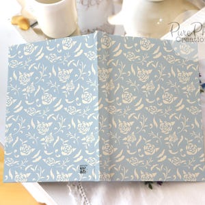 May include: A light blue notebook with a white floral pattern. The notebook is closed and lying flat on a white surface.