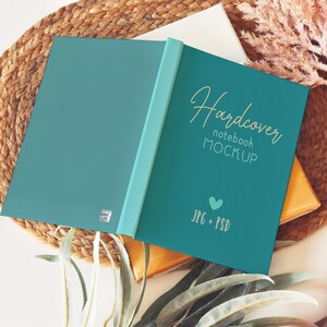Pode incluir: Maquete de caderno de capa dura teal, aberto. A capa apresenta o texto dourado "Hardcover notebook MOCKUP", um cora&ccedil;&atilde;o e "JPG + PSD". O caderno est&aacute; sobre uma superf&iacute;cie tecida.