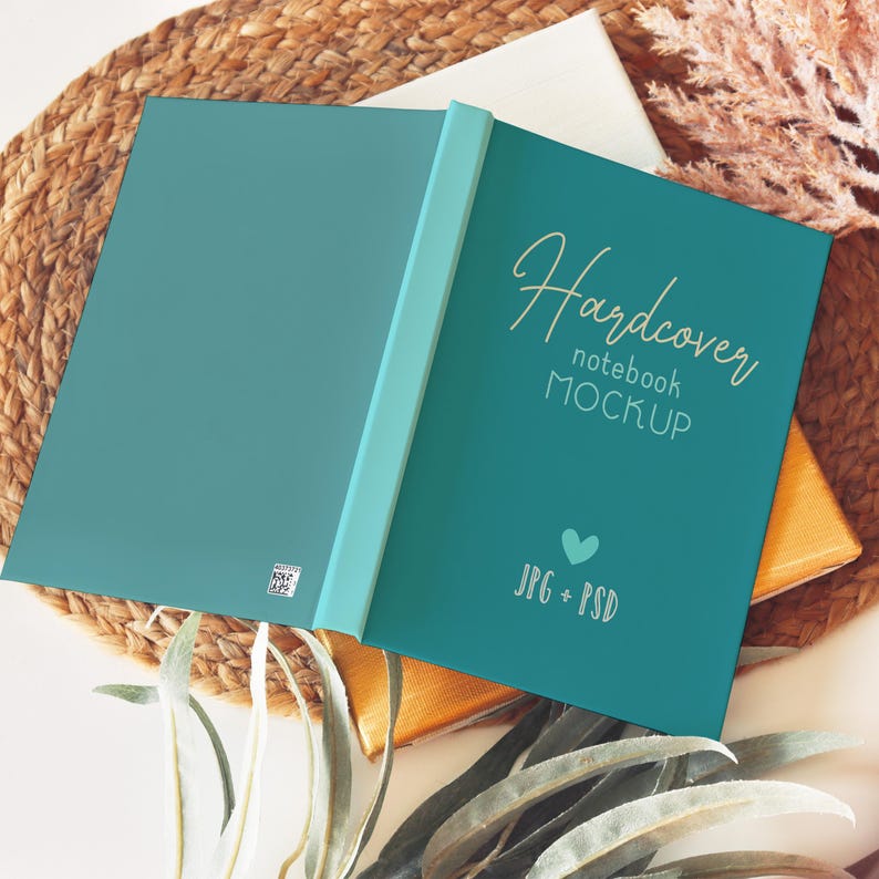 Pode incluir: Maquete de caderno de capa dura teal, aberto. A capa apresenta o texto dourado "Hardcover notebook MOCKUP", um cora&ccedil;&atilde;o e "JPG + PSD". O caderno est&aacute; sobre uma superf&iacute;cie tecida.