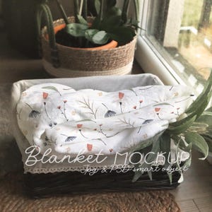 Op de afbeelding: Een wit katoenen babydeken met een bloemenpatroon in roze, blauwe en groene tinten. Het deken is gevouwen en geplaatst in een rieten mand met een kantversiering. De tekst "Blanket Mockup Jpg & PSD smart object" is zichtbaar onderaan de afbeelding.