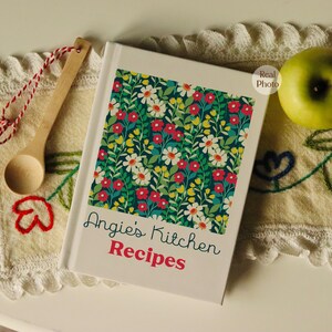 Puede incluir: Un libro de cocina blanco con un dise&ntilde;o floral en la portada. El t&iacute;tulo del libro es "Angie's Kitchen Recipes".