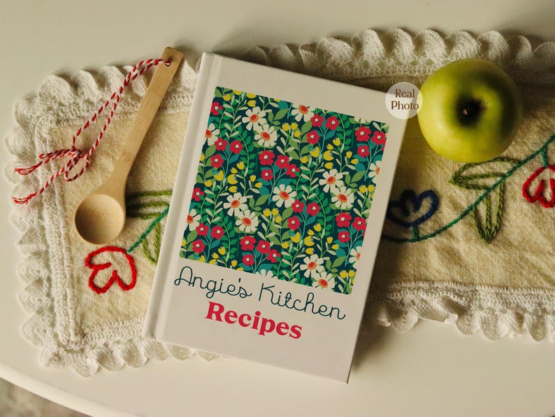Puede incluir: Un libro de cocina blanco con un dise&ntilde;o floral en la portada. El t&iacute;tulo del libro es "Angie's Kitchen Recipes".