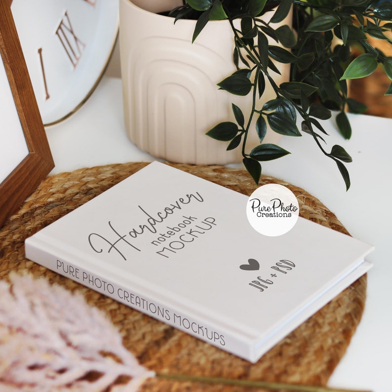 Pode incluir: Maquete de caderno de capa dura branca com o texto "Hardcover notebook MOCKUP" e "PURE PHOTO CREATIONS MOCKUPS" na capa. O caderno est&aacute; sobre um tapete tecido marrom com uma pequena planta em vaso e um rel&oacute;gio branco no fundo.