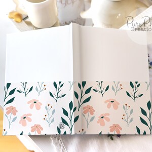 May include: A white journal with a floral design on the bottom edge. The design features pink flowers and green leaves on a white background.