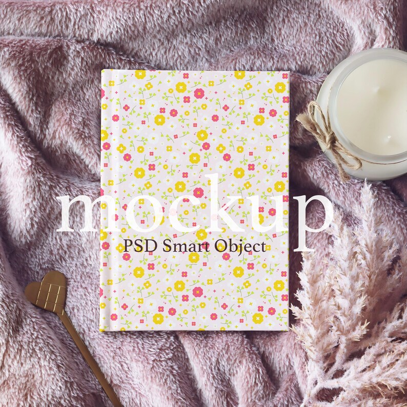 May include: A white notebook with a pink and yellow floral pattern. The notebook is lying on a pink fuzzy blanket with a candle and dried flowers.