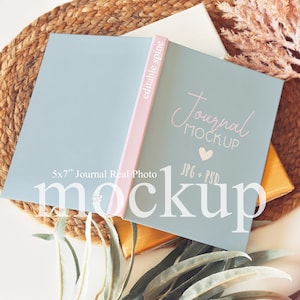 May include: An open journal mockup with a light blue cover and pink spine, featuring the text "Journal Mockup" and "5x7" Journal Real Photo". The journal rests on a woven mat with decorative elements.