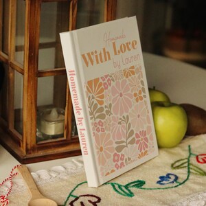 Puede incluir: Un libro de cocina blanco con un dise&ntilde;o floral en la portada. El t&iacute;tulo es "Homemade With Love by Lauren".