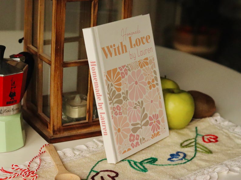 Puede incluir: Un libro de cocina blanco con un dise&ntilde;o floral en la portada. El t&iacute;tulo es "Homemade With Love by Lauren".