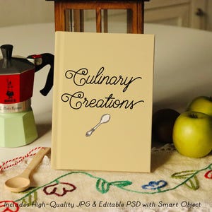 Puede incluir: Un libro beige con el texto "Culinary Creations" en letra cursiva negra. Una peque&ntilde;a cuchara de plata est&aacute; ilustrada en la portada. El libro est&aacute; sentado en una mesa blanca con un mantel a cuadros rojo y blanco.