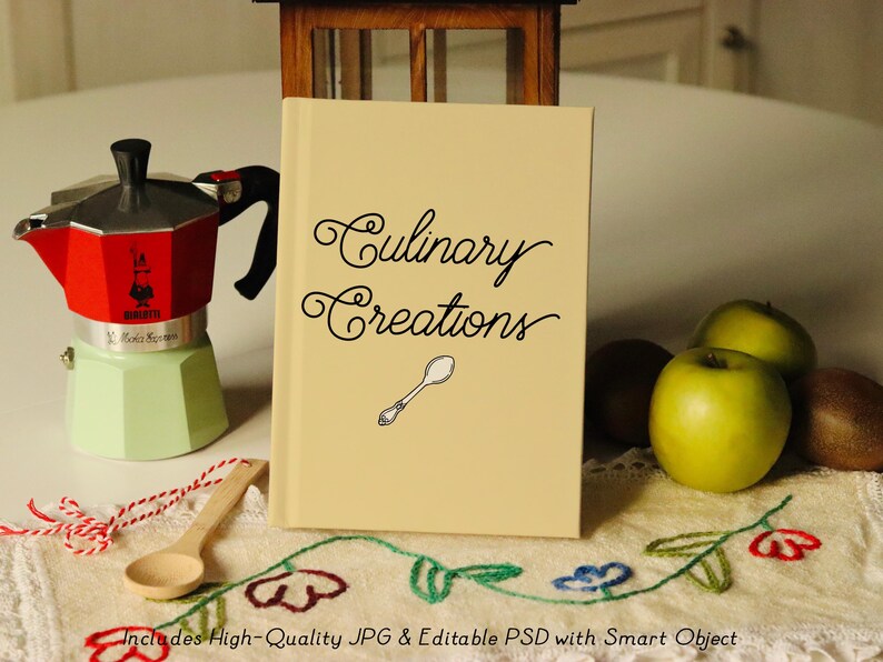 Puede incluir: Un libro beige con el texto "Culinary Creations" en letra cursiva negra. Una peque&ntilde;a cuchara de plata est&aacute; ilustrada en la portada. El libro est&aacute; sentado en una mesa blanca con un mantel a cuadros rojo y blanco.