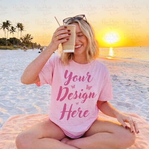 May include: A pink t-shirt with the text "Your Design Here" in a script font, worn by a person on a beach. The person is holding a coffee cup with a straw, with sunglasses on their head. The background features a sunset over the ocean.