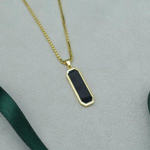 Puede incluir: Un collar dorado con un colgante rectangular negro. El colgante tiene un borde dorado y cuelga de una delicada cadena dorada. Una cinta verde oscuro está en el fondo.