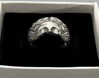 Könnte beinhalten: Ein silberfarbener Ring mit einem skulptierten Gesicht mit geschlossenen Augen und welligem Haar. Der Ring wird in einer weißen Schachtel mit schwarzem Innenraum präsentiert, die die feinen Details des Schmucks hervorhebt.