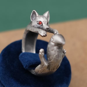 May include: A silver ring depicting a fox and a small animal. The fox has red eyes and is reaching out to the other animal. The ring is displayed on a blue velvet surface.