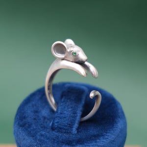 May include: A silver-toned ring featuring a mouse design. The mouse has a green gemstone eye and is perched on the ring's band. The ring is displayed on a blue velvet surface against a green background.