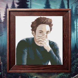 May include: A framed cross-stitch portrait of a man with dark hair and a blue sweater. He is looking down and has his hand covering his mouth.