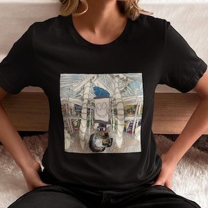 Puede incluir: Camiseta negra con una colorida impresión gráfica. La impresión muestra una ilustración detallada de un edificio con una cara sonriente y un coche. La obra de arte está en un estilo artístico único.