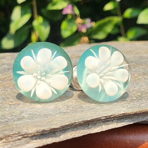 May include: A pair of teal blue glass cabochons with white flower designs. The cabochons are round and have a smooth, glossy finish.