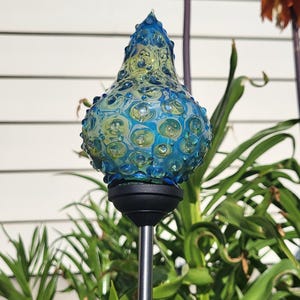 Könnte beinhalten: Dekorativer Gartenstecker mit blau-gelbem Glasaufsatz. Das Glas hat ein strukturiertes, blasenartiges Design. Der Stecker ist auf einem schwarzen Sockel und einer silbernen Stange montiert. Grüne Pflanzen und orangefarbene Blumen im Hintergrund.