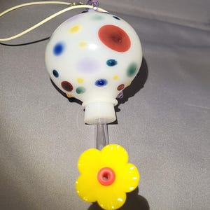 Könnte beinhalten: Ein weißer Glasflaschenverschluss mit bunten Tupfen und einer gelben blütenförmigen Basis. Der Verschluss hat ein klares Kunststoffrohr und eine cremefarbene Schnur zum Aufhängen.