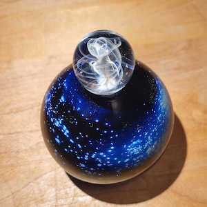 May include: A glass paperweight featuring a dark blue base with white speckles, resembling a starry night sky. A smaller clear glass orb with swirling white patterns sits atop the base, creating a unique decorative piece.