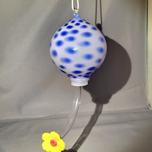 Könnte beinhalten: Ein mundgeblasener Glas-Kolibri-Futterautomat mit einer weiß-blauen, gepunkteten Kugel. Ein klarer Kunststoffschlauch erstreckt sich von unten, und eine gelbe Blume mit roter Mitte ist befestigt. Oben ist eine weiße Schnur zum Aufhängen befestigt.
