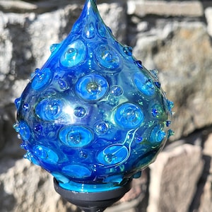 Könnte beinhalten: Ein blauer Gartenstecker aus Glas mit tropfenförmiger Spitze. Das Glas ist tiefblau mit helleren blauen und grünen Akzenten und hat eine strukturierte Oberfläche mit blasenartigen Details. Der Stecker ist auf einem schwarzen Sockel und einem silbernen Pfahl montiert.
